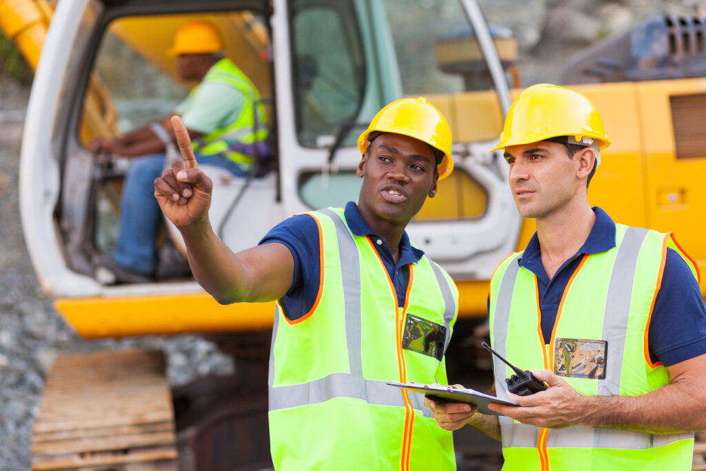 An African-American man and Caucasian man in front of heavy equipment managing a construction project.